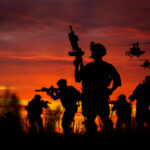 Silhouetted soldiers and helicopters against a sunset sky.