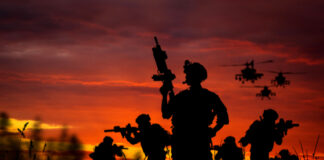 Silhouetted soldiers and helicopters against a sunset sky.