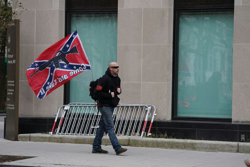 shutterstock_1888462168.jpg Man walking with Confederate flag on city sidewalk.