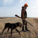 Ballot SHOCKER: Hunting & Fishing Under Fire…. A man in hunting gear walking with a dog in a field during sunset