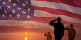 Silhouetted soldiers saluting against an American flag backdrop during sunset
