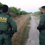 Two U.S. Border Patrol officers standing on a dirt path.
