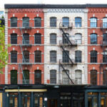 Red-brick building with fire escapes and storefronts, daytime.